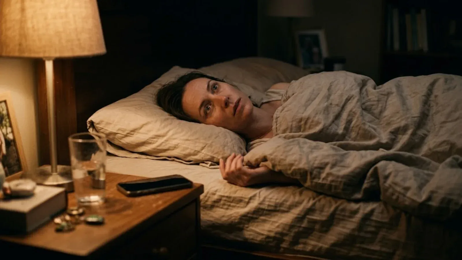A woman lying awake in bed at night beside a dim bedside lamp, phone face-down on the nightstand
